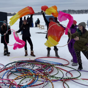 A group of performers wave colorful fabric in the air. A huge pile of hula hoops rests in front of them on the frozen lake.