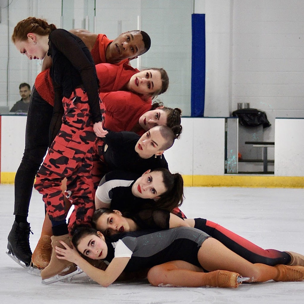 A group of skaters at an indoor rink pose in a sculptural and dynamic pile