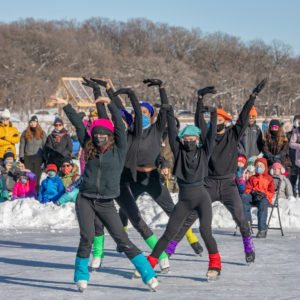 A group of ice skaters wearing black with colorful legwarmers and berets pose mid-performance with onlookers behind them