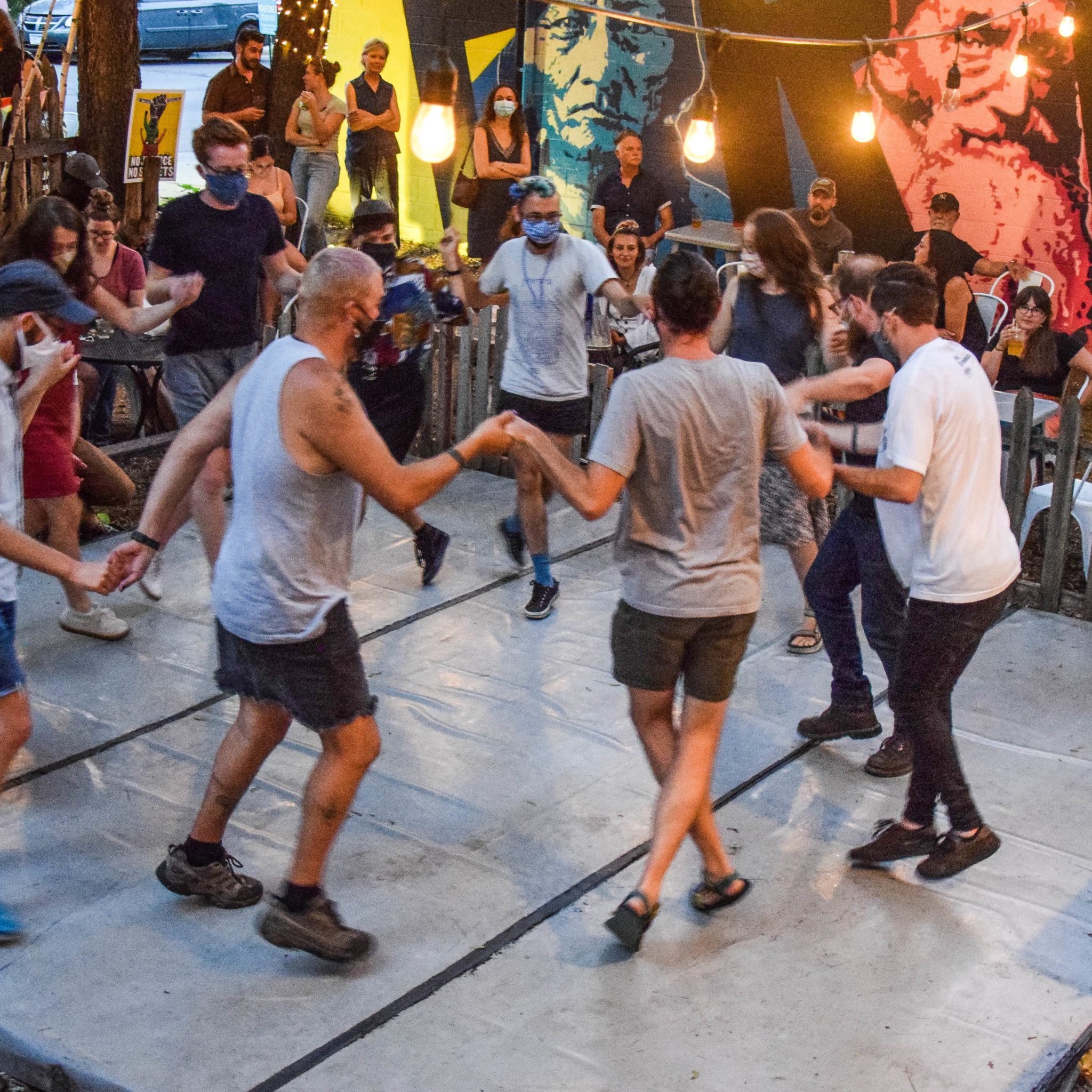 A group of people in summer clothes dance in a circle while holding hands. A crowd watches.