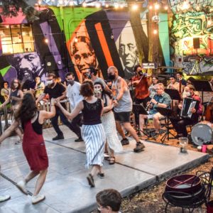 Live musicians play for a group of people dancing in a circle. Big colorful murals are in the background of the outdoor scene.
