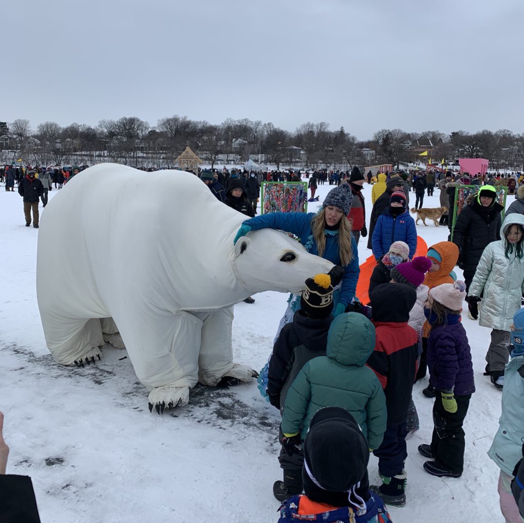 a life size polar bear puppet is swarmed by kids in snowsuits on a frozen lake.