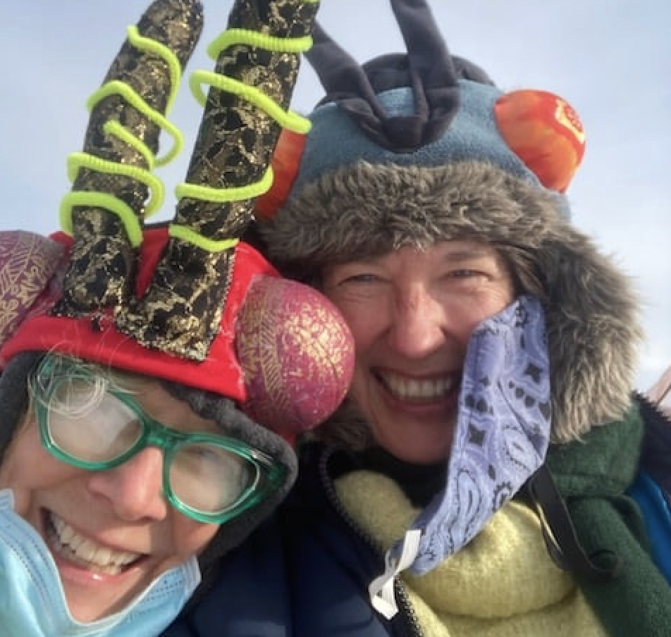 A closeup of two people wearing pollinator hats and antennae smiling in the cold.
