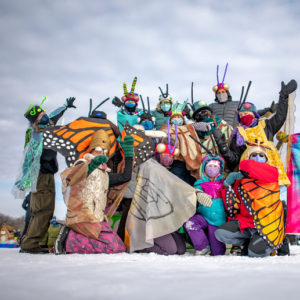 A group of adults pose in their vibrant pollinator costumes on the frozen lake.