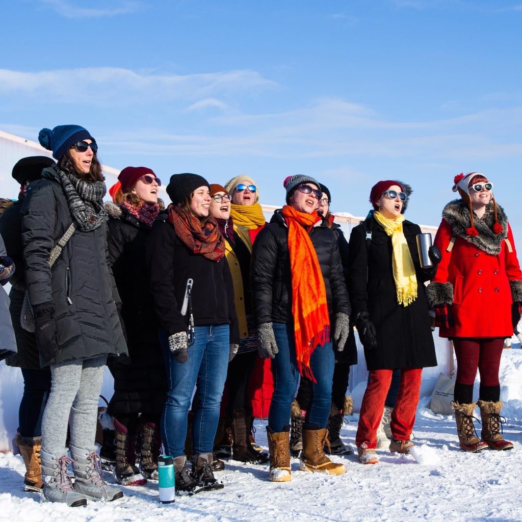 A group of singers in choir formation sing on a frozen lake against a light blue sky