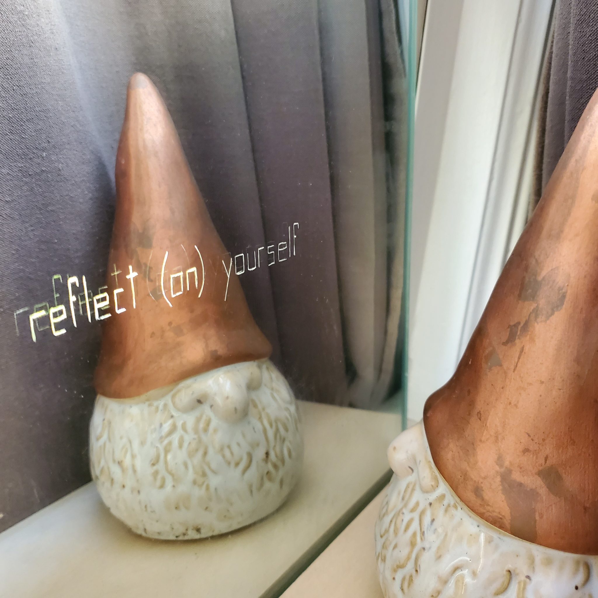 A gnome figure with a bronze pointy hat looks at themself in the mirror. Words on their reflection read 'reflect (on) yourself'