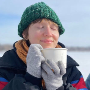 A person bundled up in multi-colored winterwear holds a cup of soup near their face in gloved hands. With eyes closed they savor the aroma of the soup.