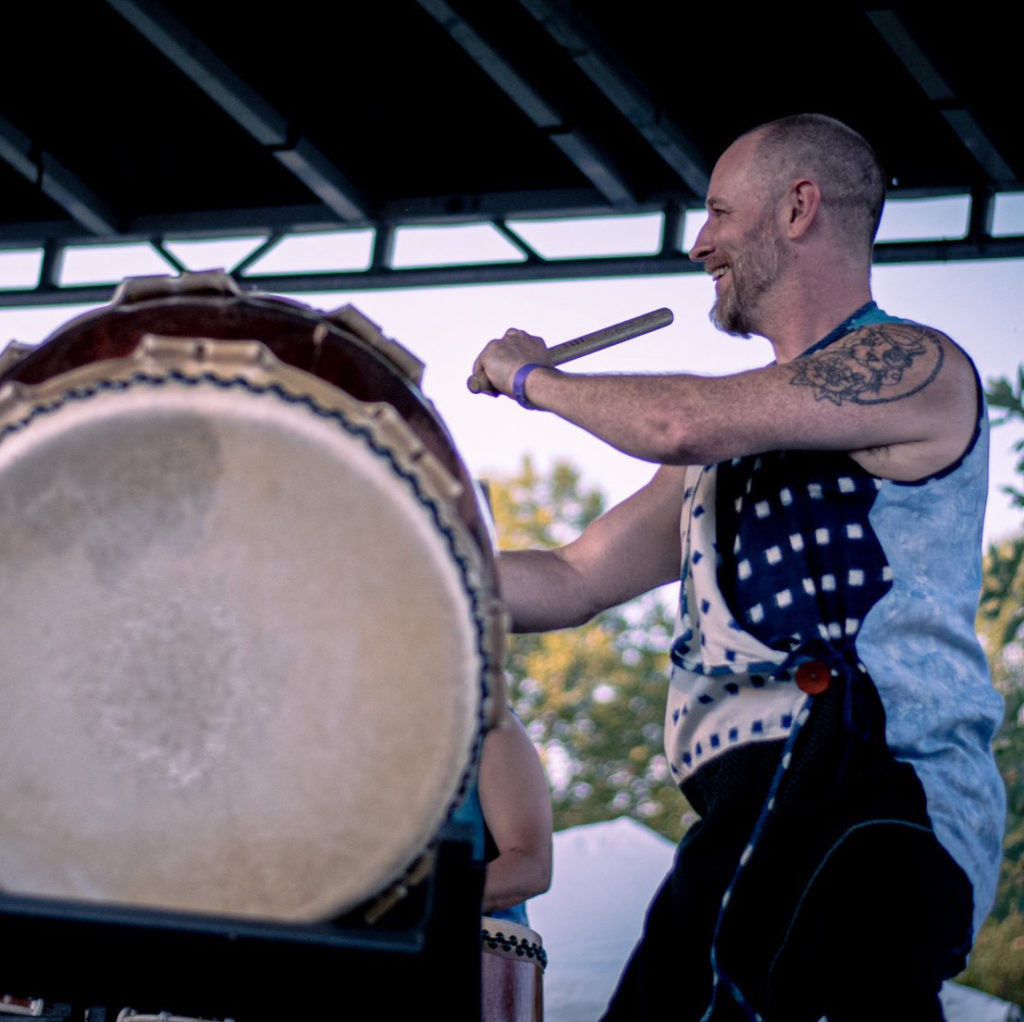 A bald person smiles as they hit a large drum