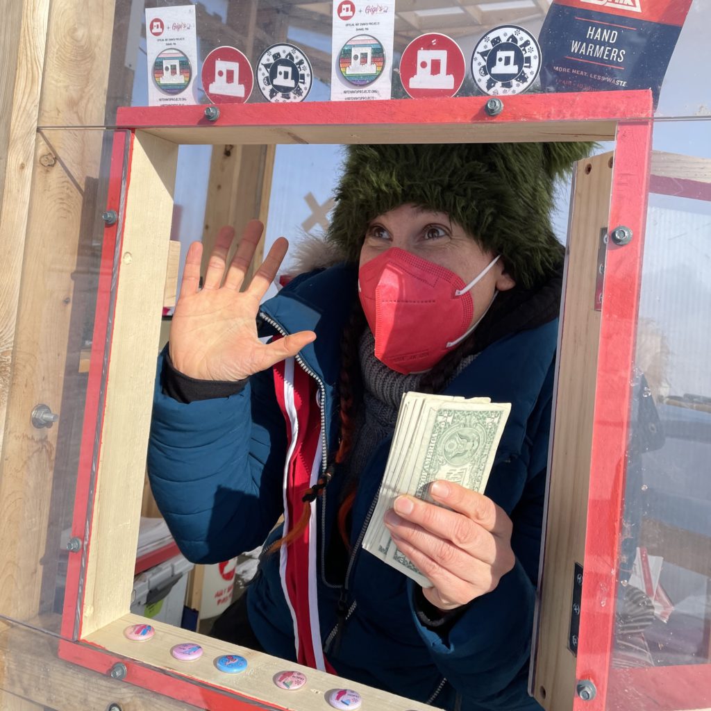 A person holding dollar bills waves with their other hand as they look out of a little porthole window of a shanty. Merch is displayed around them.