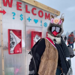 A person in a shantastic getup of fur, goggles, flowers and such, stands in front of the welcome shanty