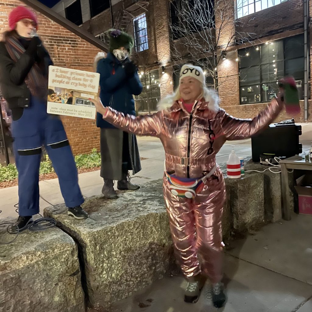 a person in a shiny pink snowsuit holds their arms out in excitement while two people stand on a ledge announcing something into a microphone.