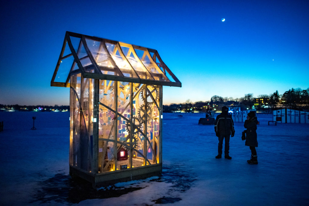 A clear shanty is lit up at night against deep blue ice and sky.