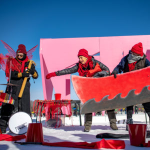 A play in action on the frozen lake! Performers are dressed in black and red against a pink backdrop shanty.