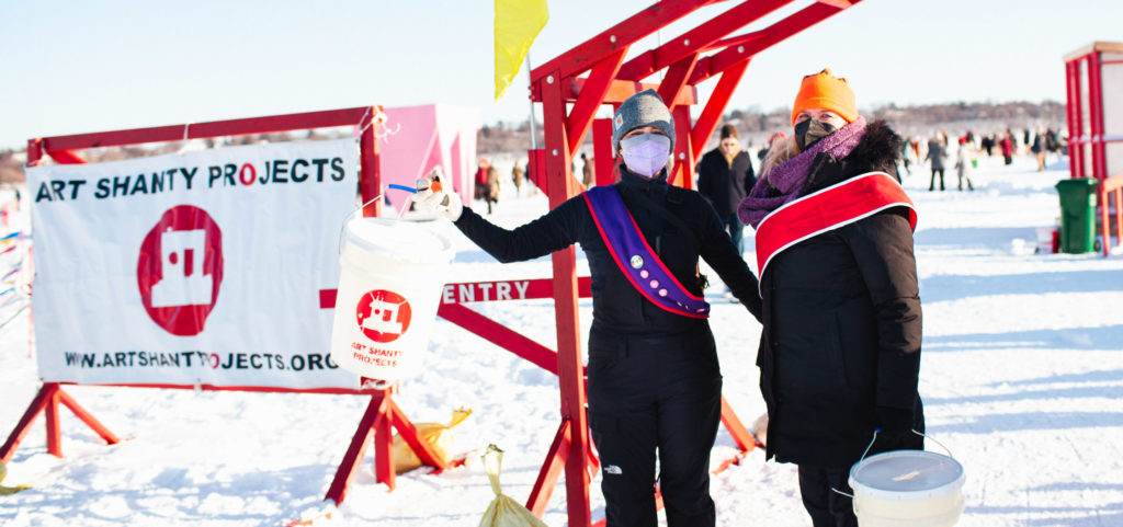 Two people in snowsuits and sashes stand under the red entrance gate and next to the Art Shanty banner.