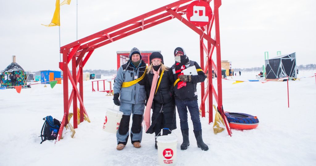 Three volunteers in sashes stand under the red entrance arch with smiles on their faces.