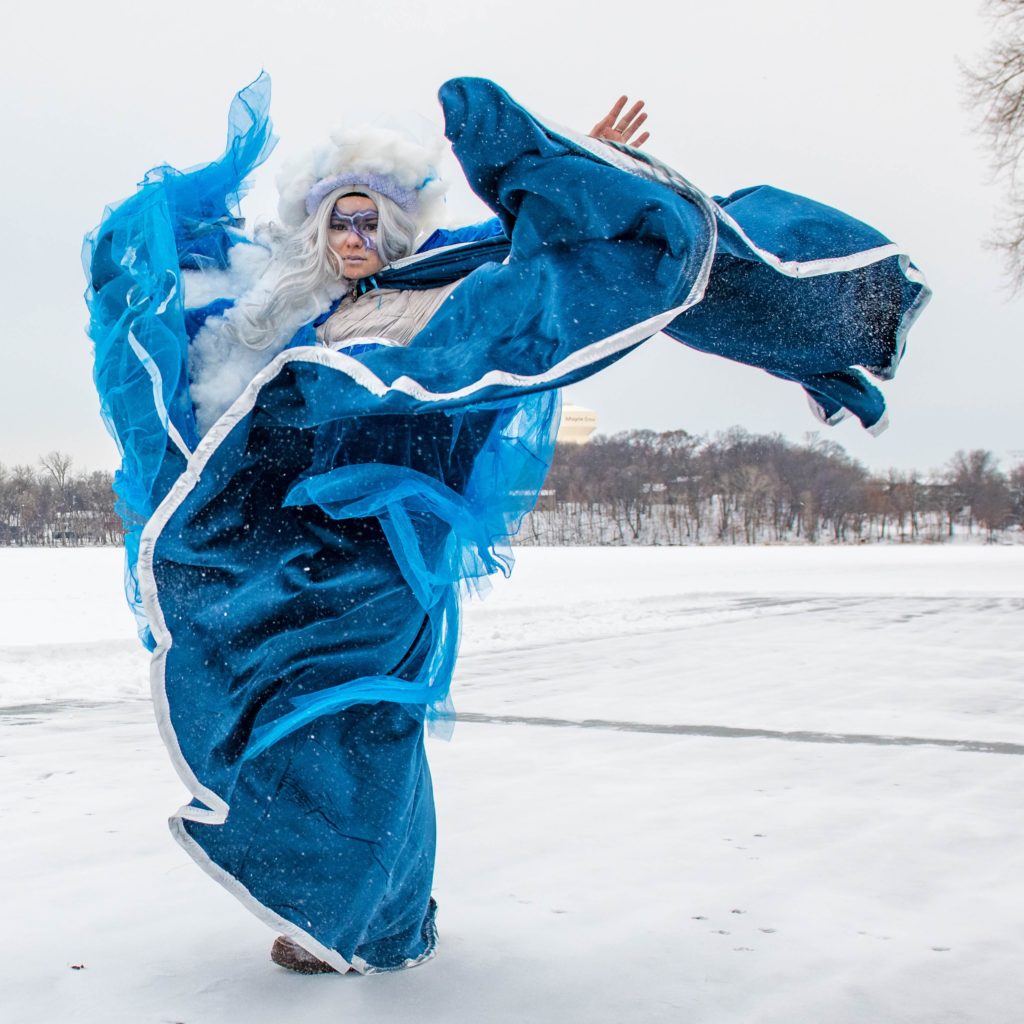 A person in a blue costume that resembles water whips the fabric in the wind on a frozen lake to imitate a storm.