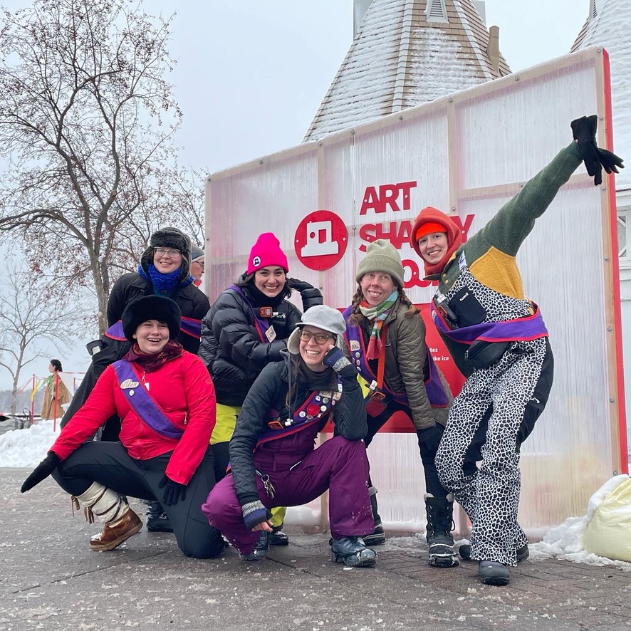 Six staff members wearing colorful winterwear strike a dynamic post outside in front of an Art Shanty sign