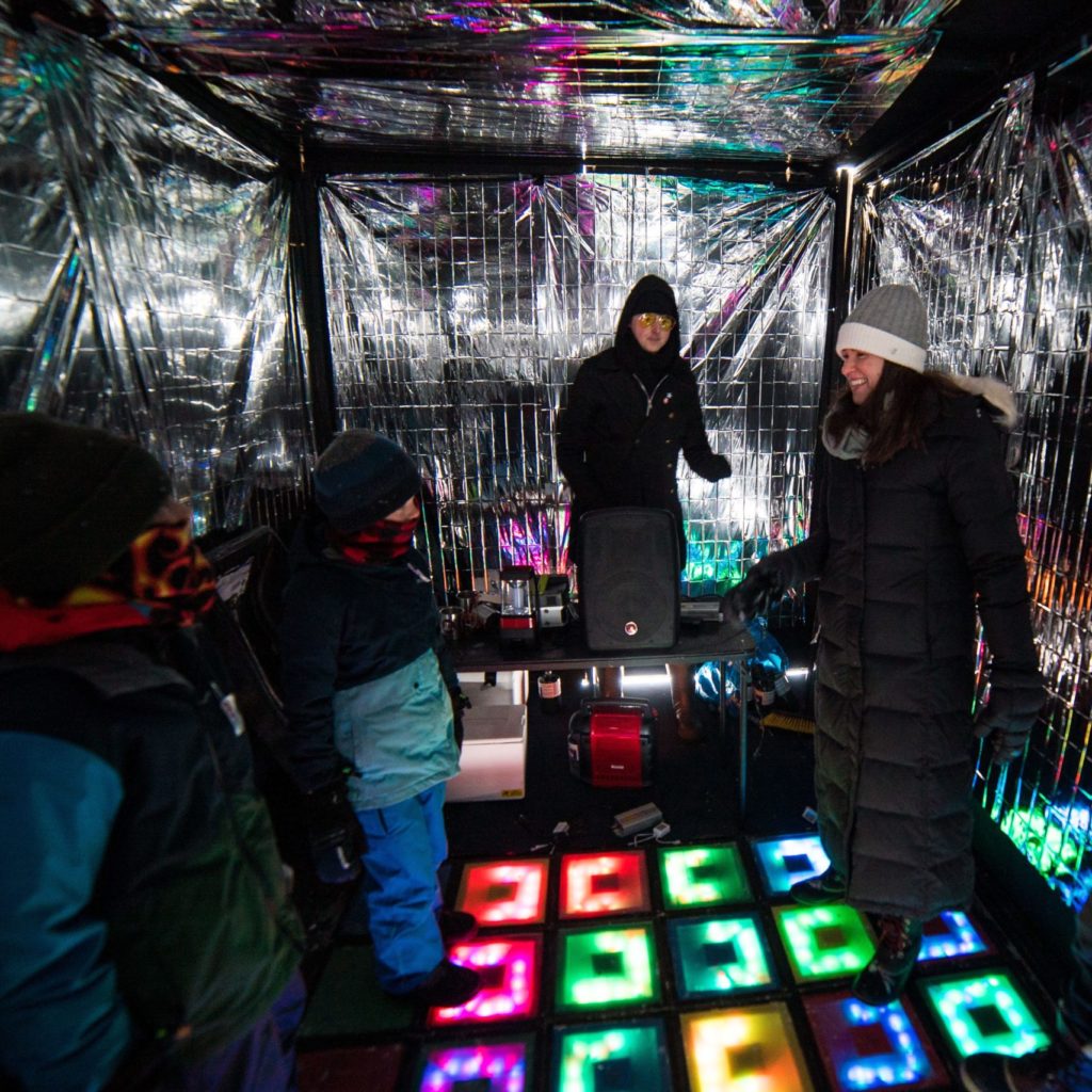 people dance inside a dark shanty with a colorful lit-up dance floor and silver space blanket walls