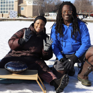 Two dark-skinned people kneel on the ice, smiling, with the bandshell in teh background