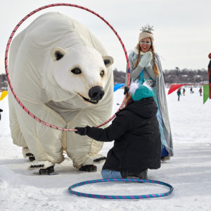 A person kneels on the snow, holding a hula hoop in front of a life sized polar bear puppet