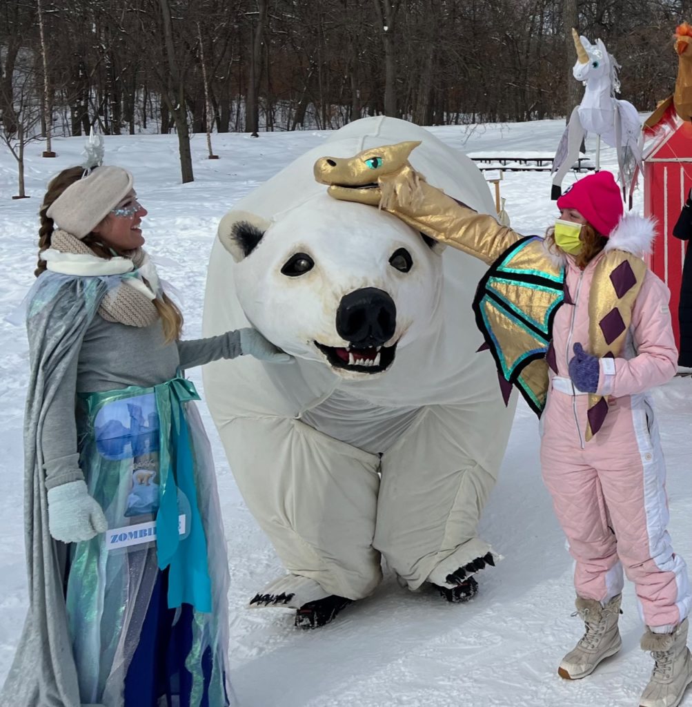 A dragon puppet pets a life sized polar bear puppet while humans watch