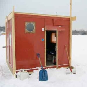a boxy red shanty on a snowy lake with a shovel resting next to the open door.