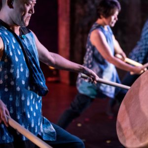 Two people dressed in blue play taiko drums with large sticks