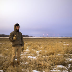 A bundled up smiling person stands in a snowy field with a pale sky in the distance