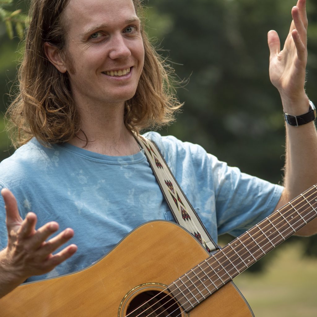 A headshot of a smiling person with long blond hair holding a guitar