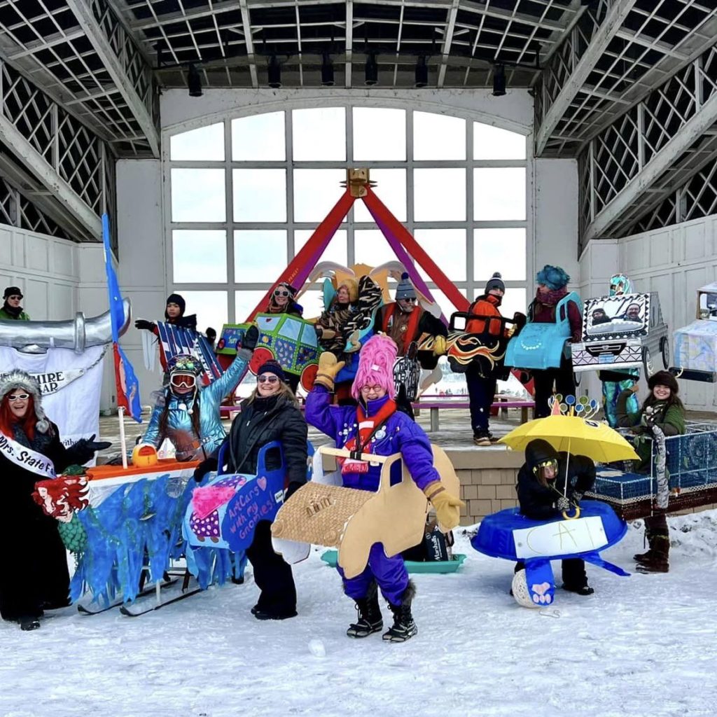 A group of adults and kids pose wearing their colorful art cars made of cardboard in front of a pyramid shaped shanty