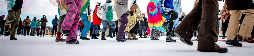 a closeup of boot-clad feet dancing on a frozen lake with spectators watching in the distance