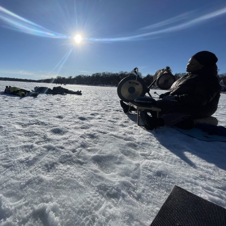 A person plays a gong while people lay on the frozen lake