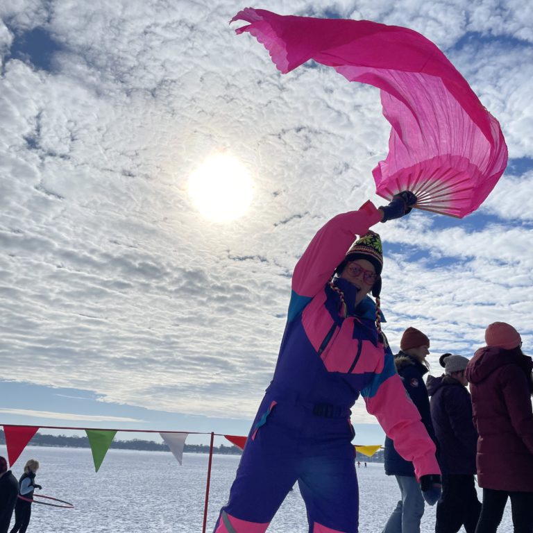 A person in a retro 80s snowsuit plays with a big pink fan against a snowy lake and cloud filled sky