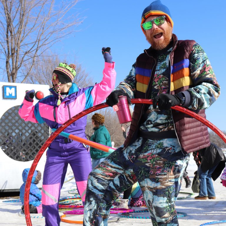 Two people in fabulous ski suits play with hula hoops. A boom box shaped shanty is in the background