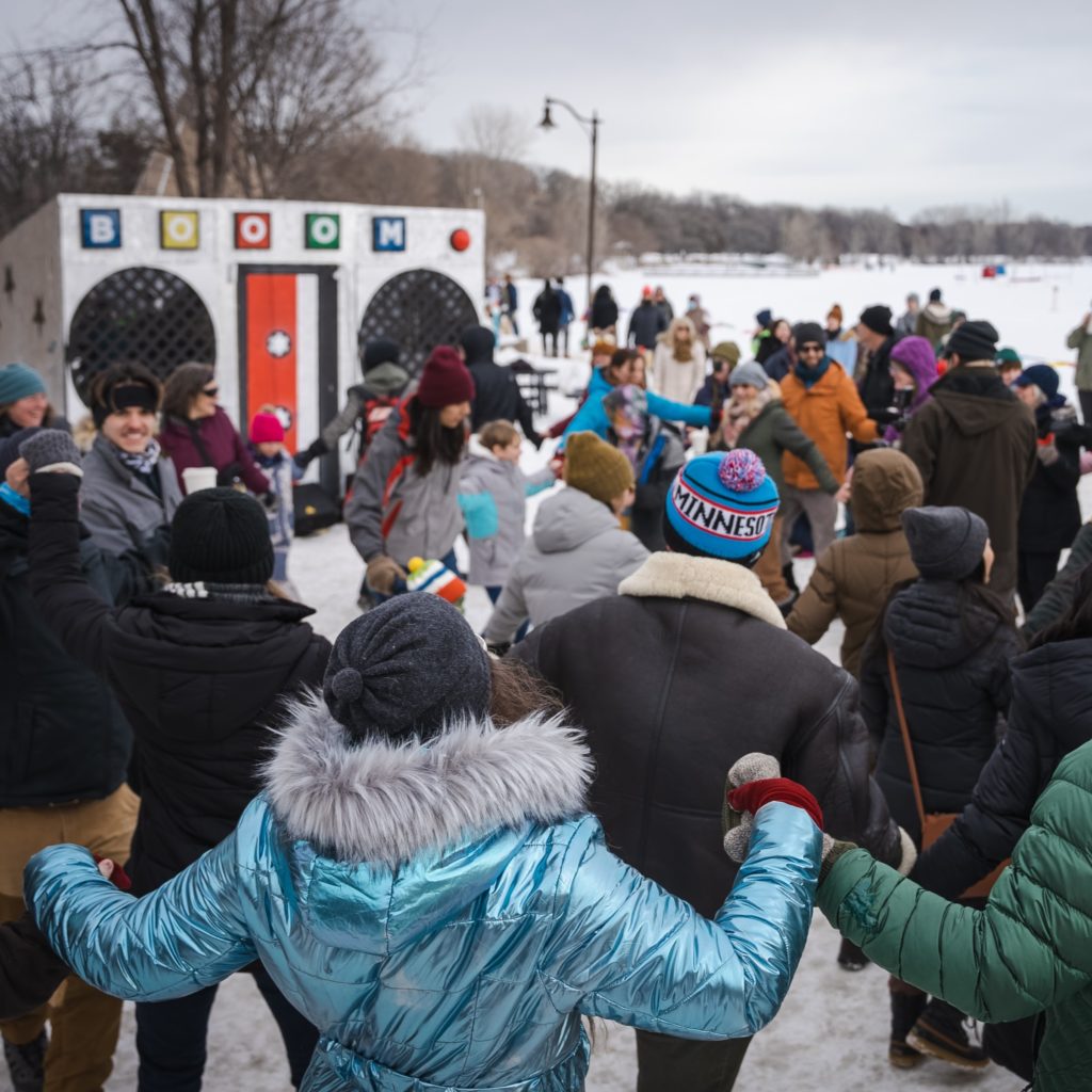 A group of people in snowsuits dance together in front of a boom box shaped shanty