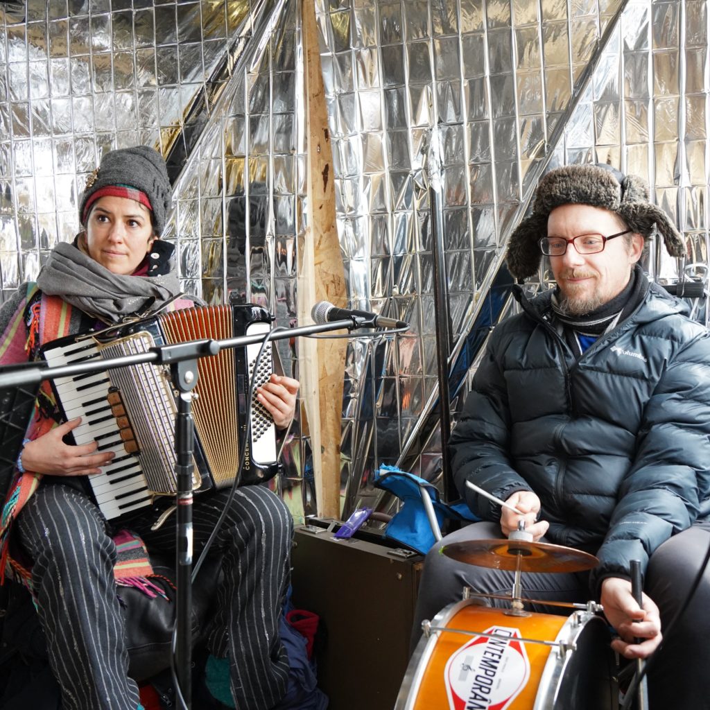 Two people play an accordian and drum against a shiny silver shanty interior