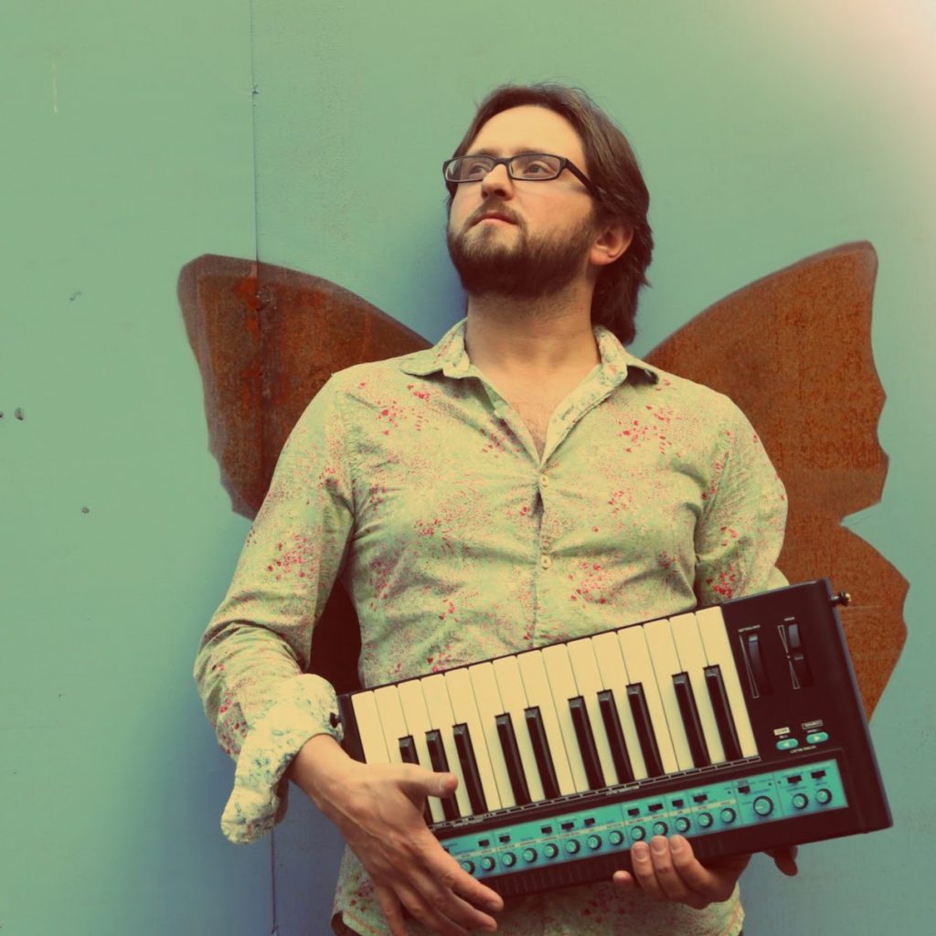 A person holds a small keyboard instrument while standing in front of a mural, appearing that they have wings