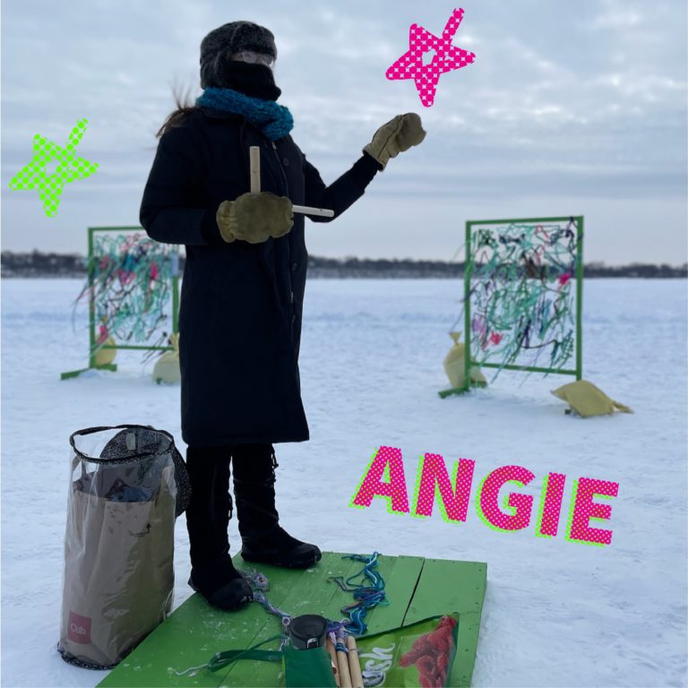 A photo of a person so bundled up you can only see their bespectacled eyes. They stand in front of a weaving installation.