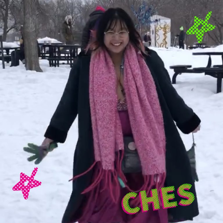 a person wearing a black coat with pink winter accessories smiles as they encounter the shanty village in a snowy landscape.