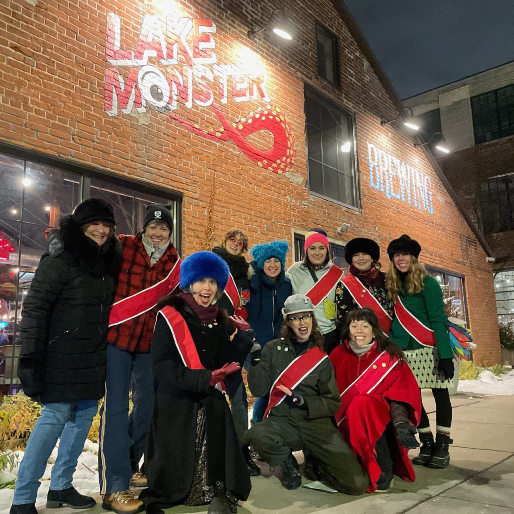 A group of people wearing winterwear and matching red sashes pose in front of Lake Monster Brewing at night