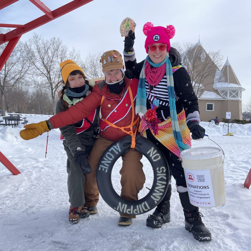 3 board members smiling and posing while collecting donations in winter gear carrying a bucket, and a floatation ring saying "no skinny dipping".
