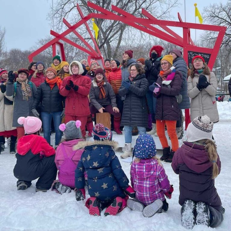A choir dressed in fiery winterwear sings on a red open air stage while a small group of kiddos eagerly listen and watch