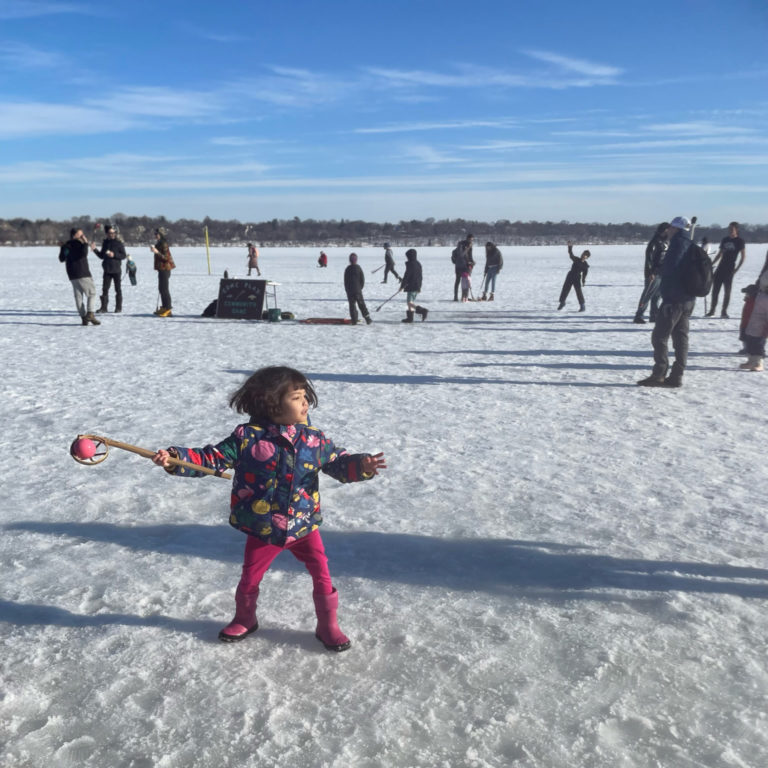A small child gets ready to toss a ball with a wooden lacrosse stick while others play in the distance on the frozen lake