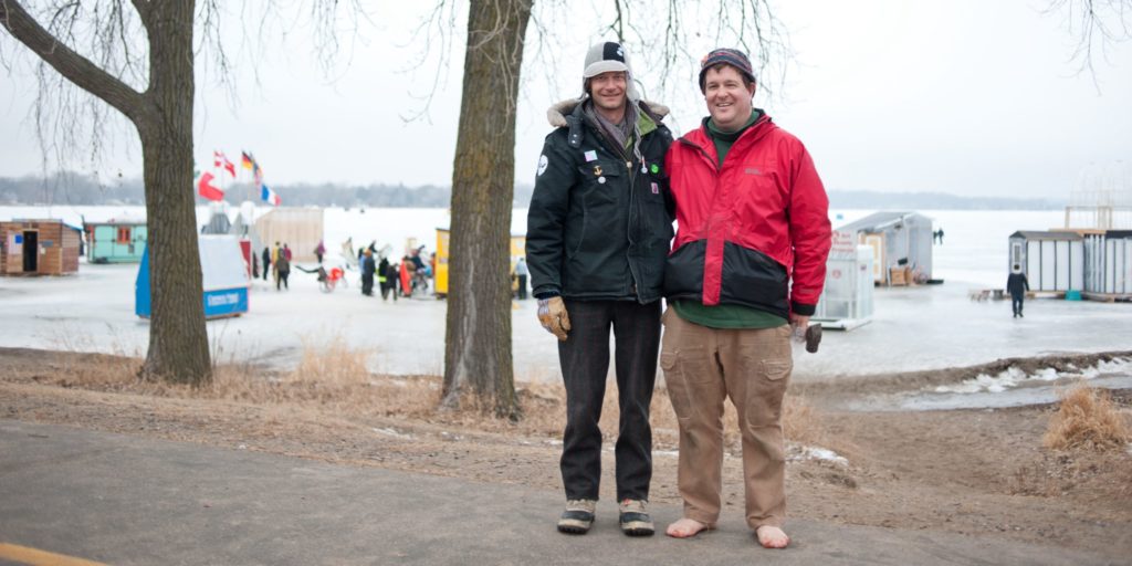 Two men stand on a path by Medicine Lake. One wears dark winter gear, the other wears a red jacket and has bare feet.