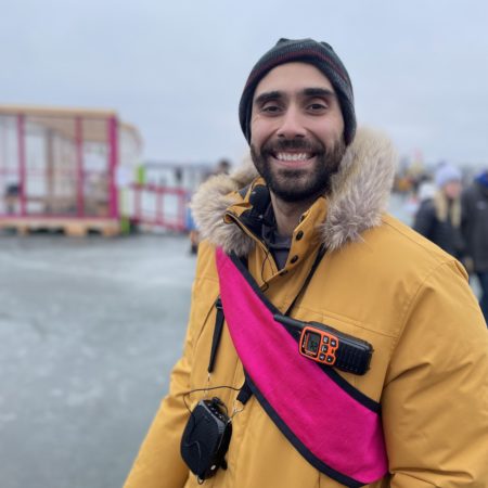 a person with a dark beard smiles on a frozen lake. he wears a yellow coat and pink sash denoting he is a board member.