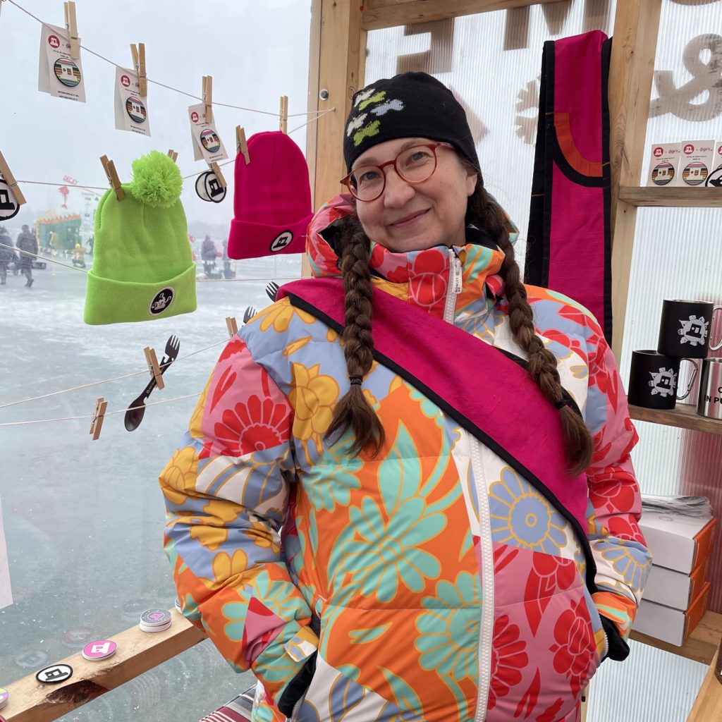 a person with long braids wears a very colorful snowsuit. a merch display of winter hats and postcards are strung across a line in the background.