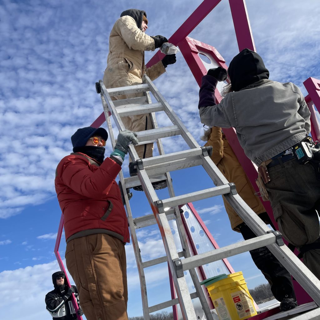 A person is at the top of a ladder while others hold shanty pieces that they're all assembling.