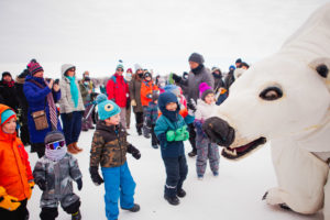 A crowd of kids in colorful snowsuits are greeted by a life size polar bear puppet on the frozen lake.