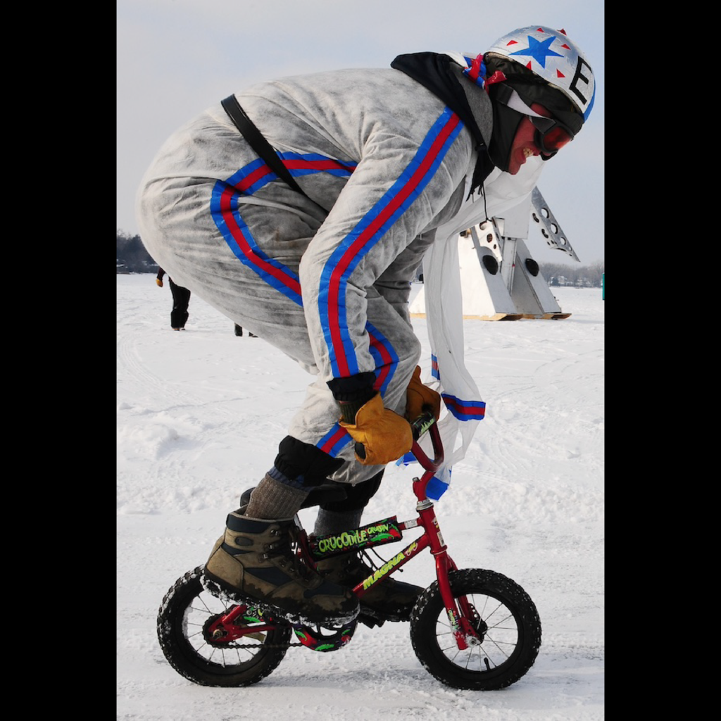 A very tall person in a racing snowsuit rides a tiny bicycle on the frozen lake