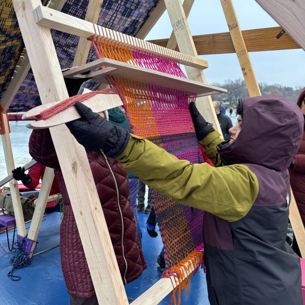 A person in winterwear reaches up to a wall panel of a shanty to weave on a giant loom